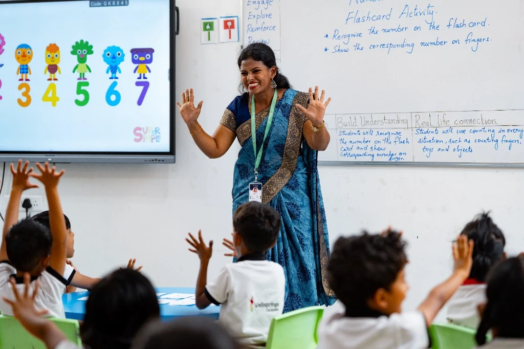 A teacher in a blue sari leading a flashcard activity, both hands raised